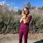 Female with long dark blonde hair wearing maroon colored scrubs holding a tabby cat