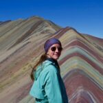 Female standing in front of mountains wearing a light blue jacket and purple scarf on her head with sunglasses